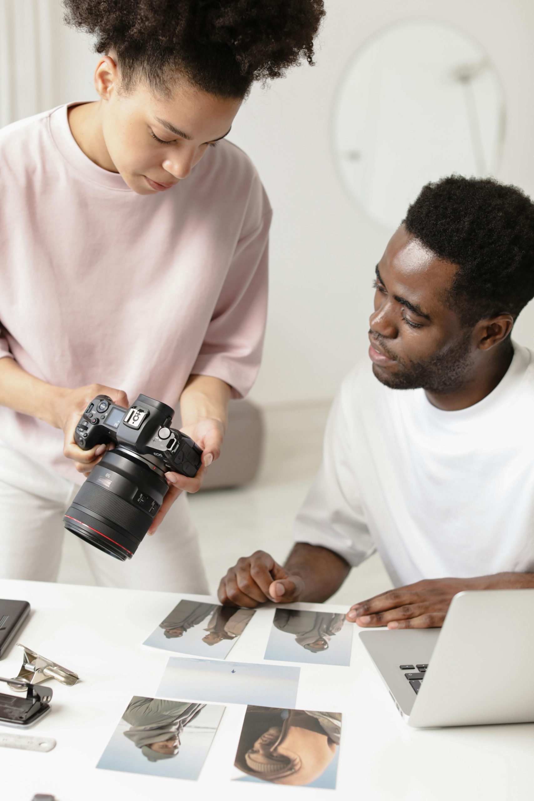 Two photographers discuss printed photographs at a table, using a DSLR and a laptop.